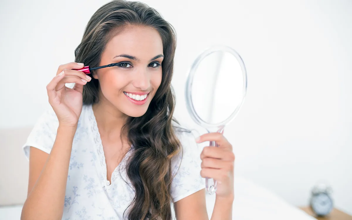 A woman smiles while holding a mirror, reflecting confidence and beauty, symbolizing self-care and eyelash enhancement tips.