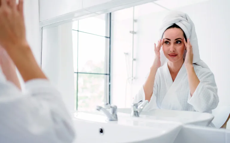 A woman in a bathrobe looks into a mirror, inspired by the skincare advantages of incorporating rice water into her routine.