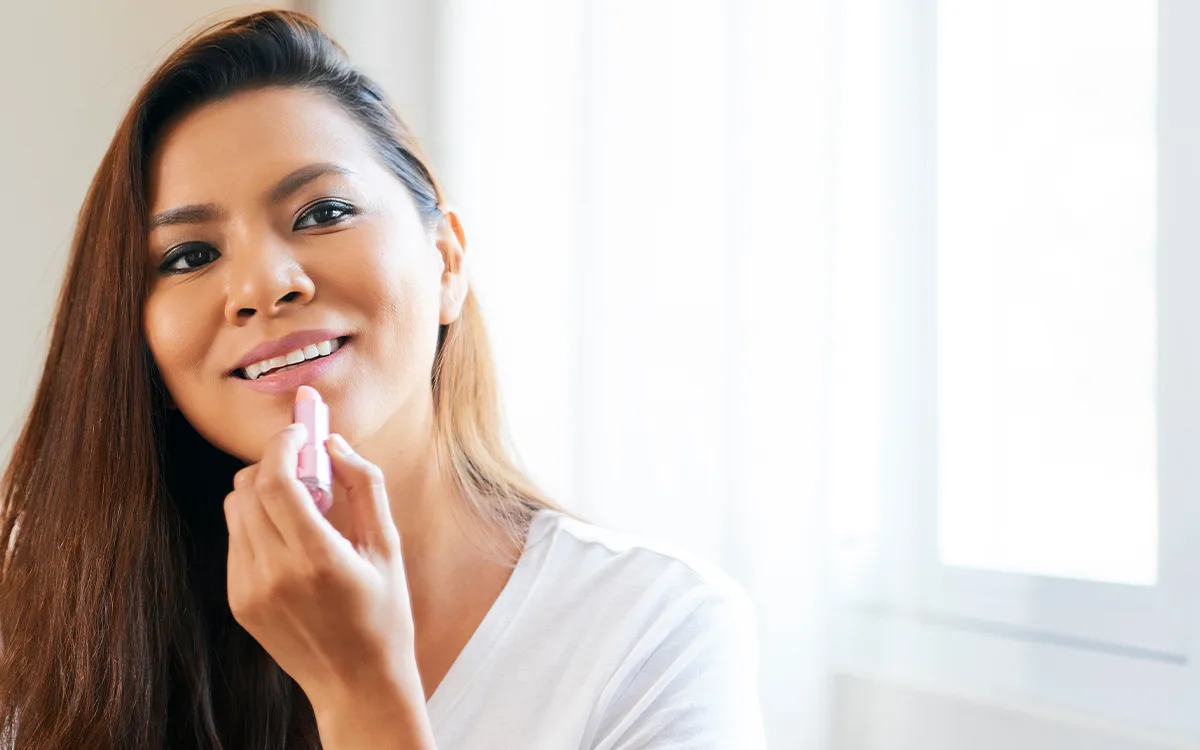 Woman applying lip balm while looking in a mirror, standing near a window.