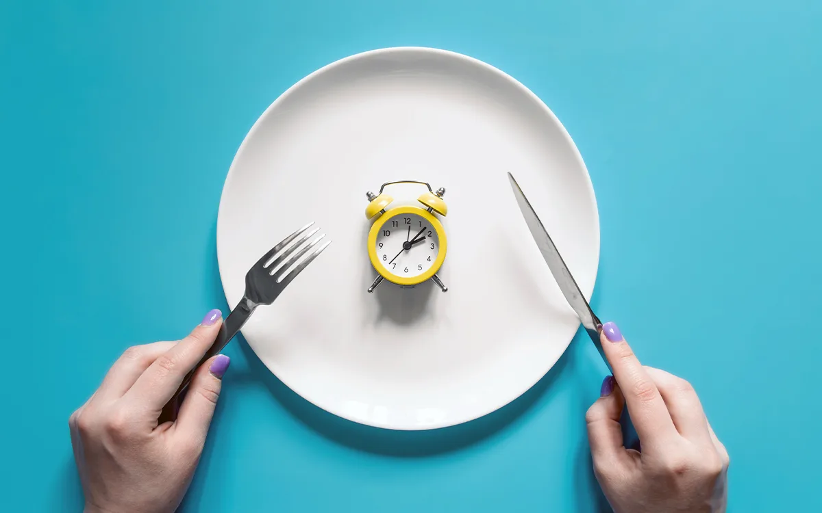A person holds a fork and knife over a plate featuring a clock, symbolizing the connection between eating and circadian rhythms.