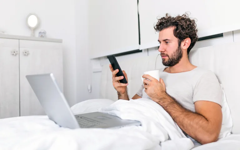 A man in bed enjoys a cup of coffee while holding a phone, embracing a tech-free morning for better focus and clarity.