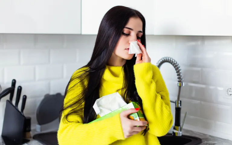 A woman blowing her nose in a kitchen, highlighting the impact of diet on immune health and overall well-being.