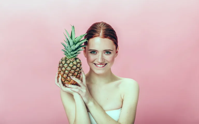 A woman holds a pineapple against a pink background, highlighting the fruit's potential skincare benefits and vibrant appeal.