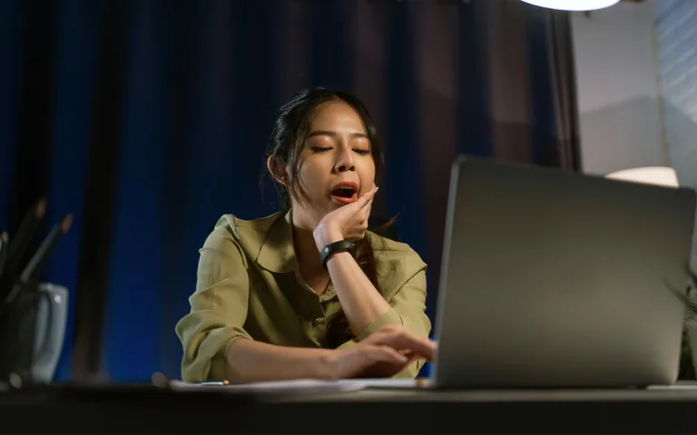 A woman at a desk, mouth open, appears tired, highlighting the struggle to stay awake during the day.