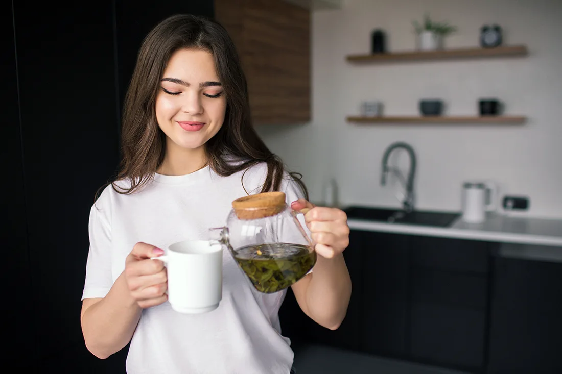 A woman holds a cup of green tea and a jug, symbolizing the tea's benefits for hair health and scalp comfort.