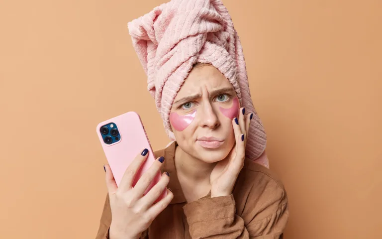 A woman with a pink towel on her head holds a pink phone, highlighting concerns about screen time and skin health.