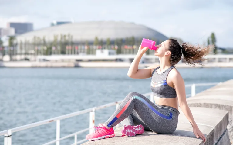 A woman in sports gear drinks from a pink water bottle, promoting hydration during outdoor activities and sun exposure.