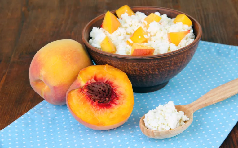 A bowl of cottage cheese topped with sliced peaches sits on a wooden table, showcasing a healthy, high-protein meal option.