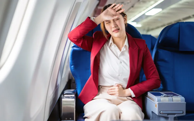 A woman on an airplane, sitting with her hands on her head, appears contemplative about travel-related digestive issues.