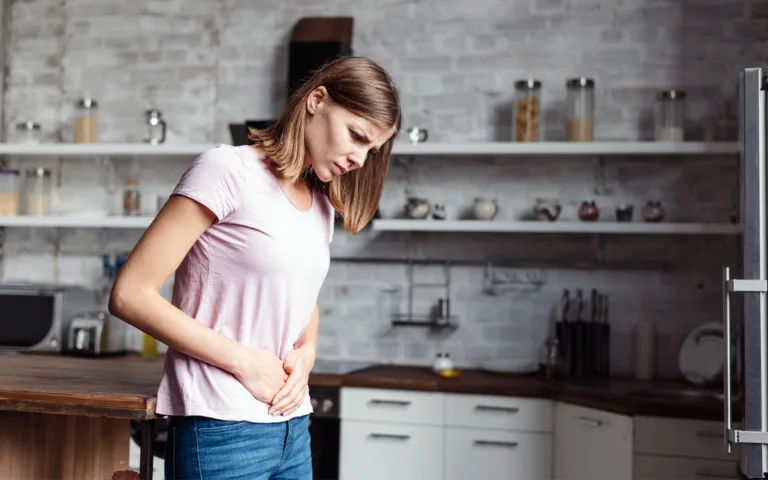 A woman in a kitchen holds her stomach, indicating discomfort, possibly due to bloating or digestive issues.