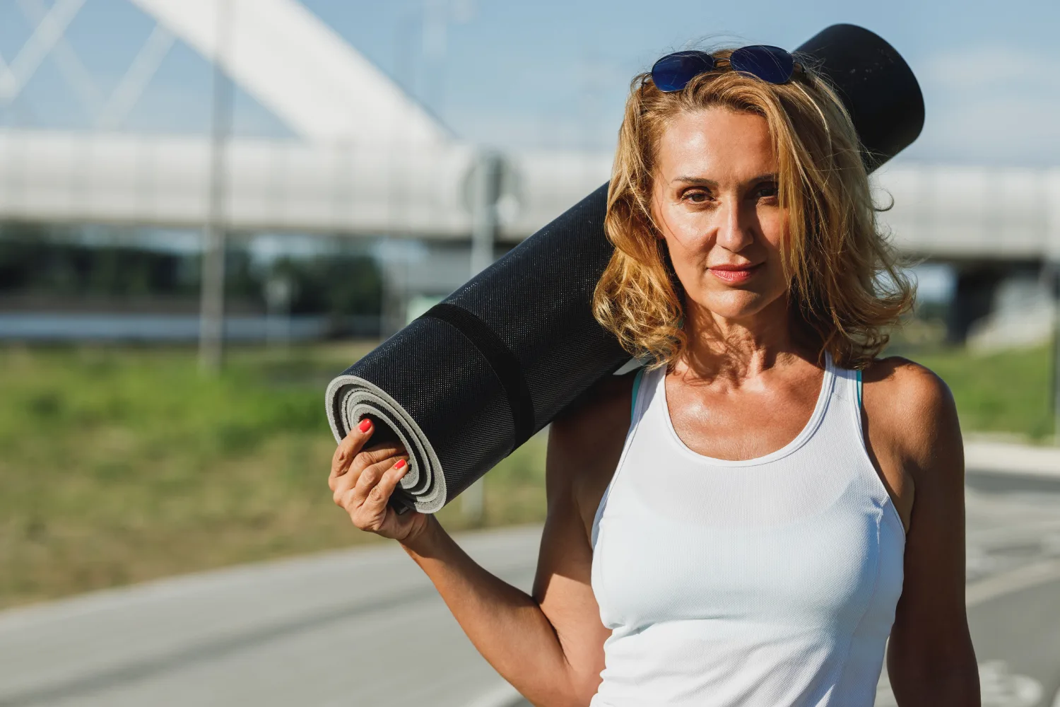 Midlife woman holding a yoga mat outdoors, representing gentle movement and regular exercise to help manage cortisol levels during menopause