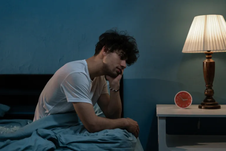 Man sitting awake on the bed at night beside a clock and lamp, illustrating insomnia and common 3 AM wake-ups.