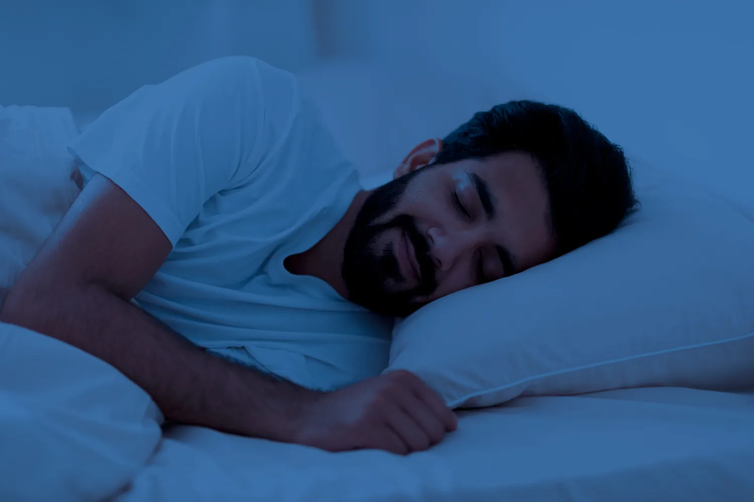 Man sleeping peacefully on his side in a dim blue-lit bedroom, symbolizing deep sleep and fewer nighttime awakenings.
