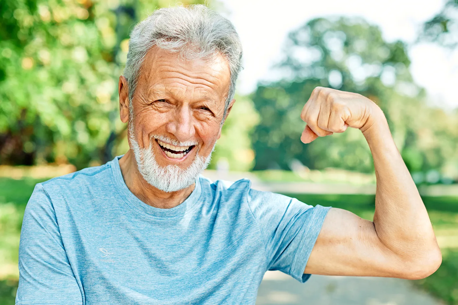 Older man smiling and flexing his arm outdoors, representing strength, healthy aging, and muscle support for seniors