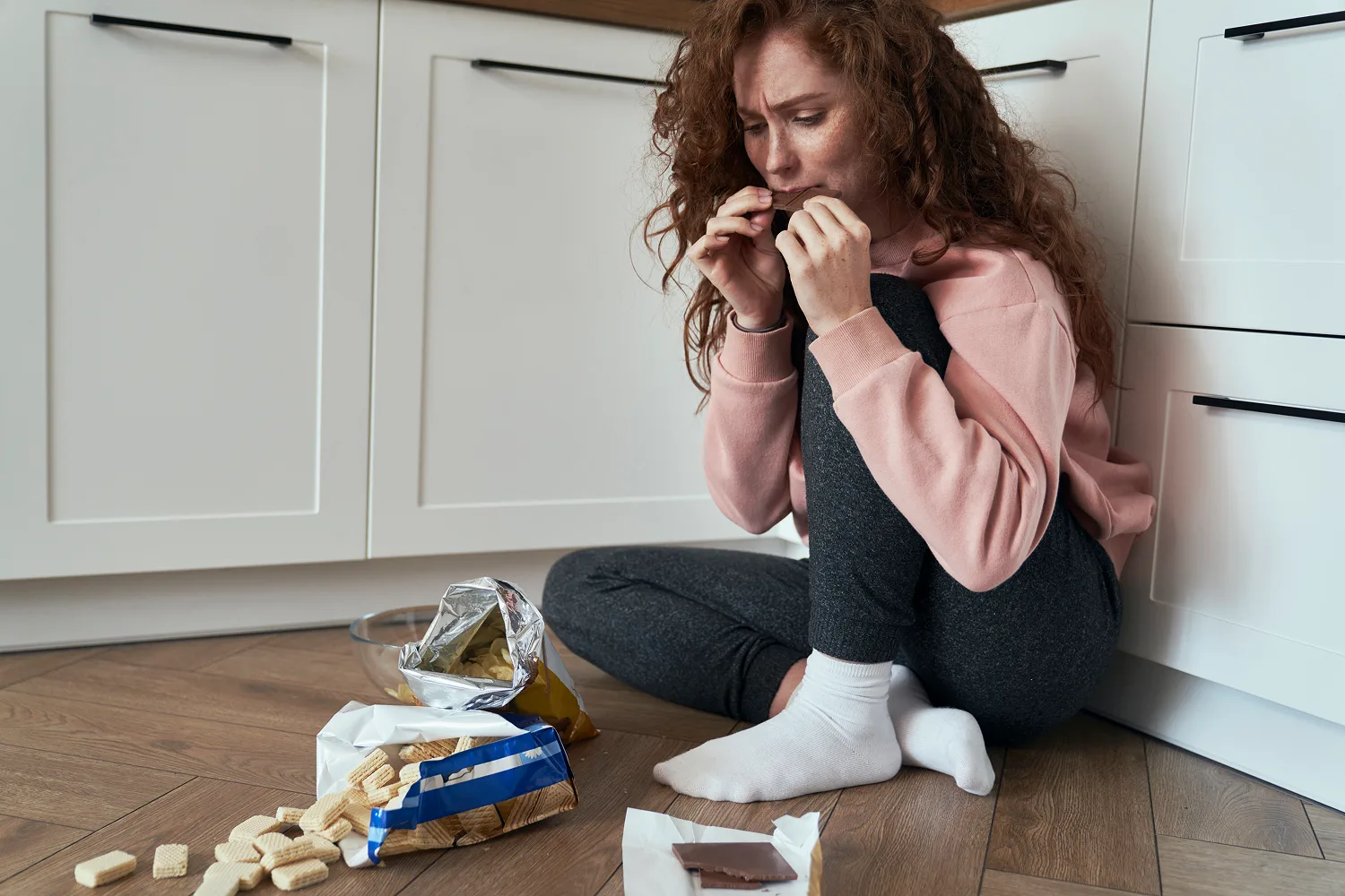 Woman stress eating snacks on kitchen floor showing emotional eating patterns linked to increased cravings and hunger